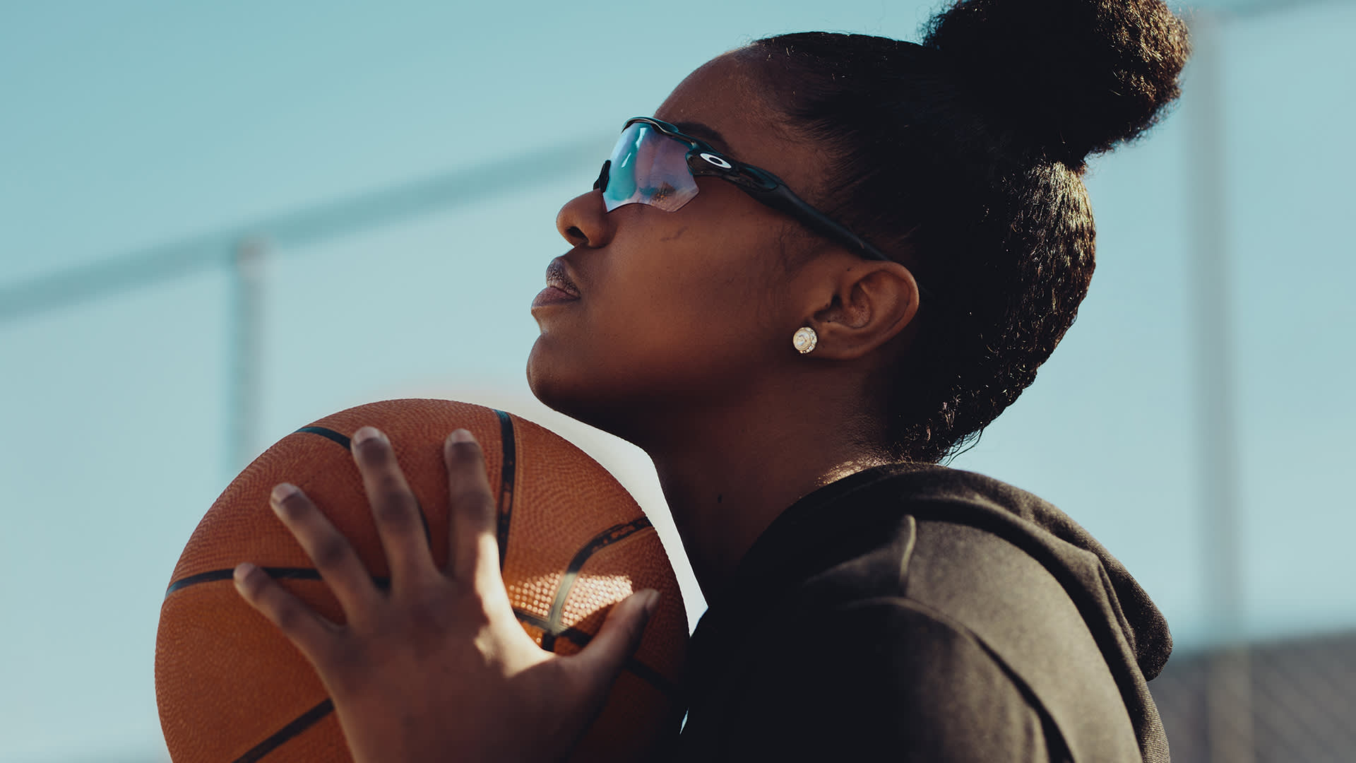 A young woman wearing sunglasses and a black hoodie holds a basketball close to her face, looking upwards with determination against a clear blue sky.