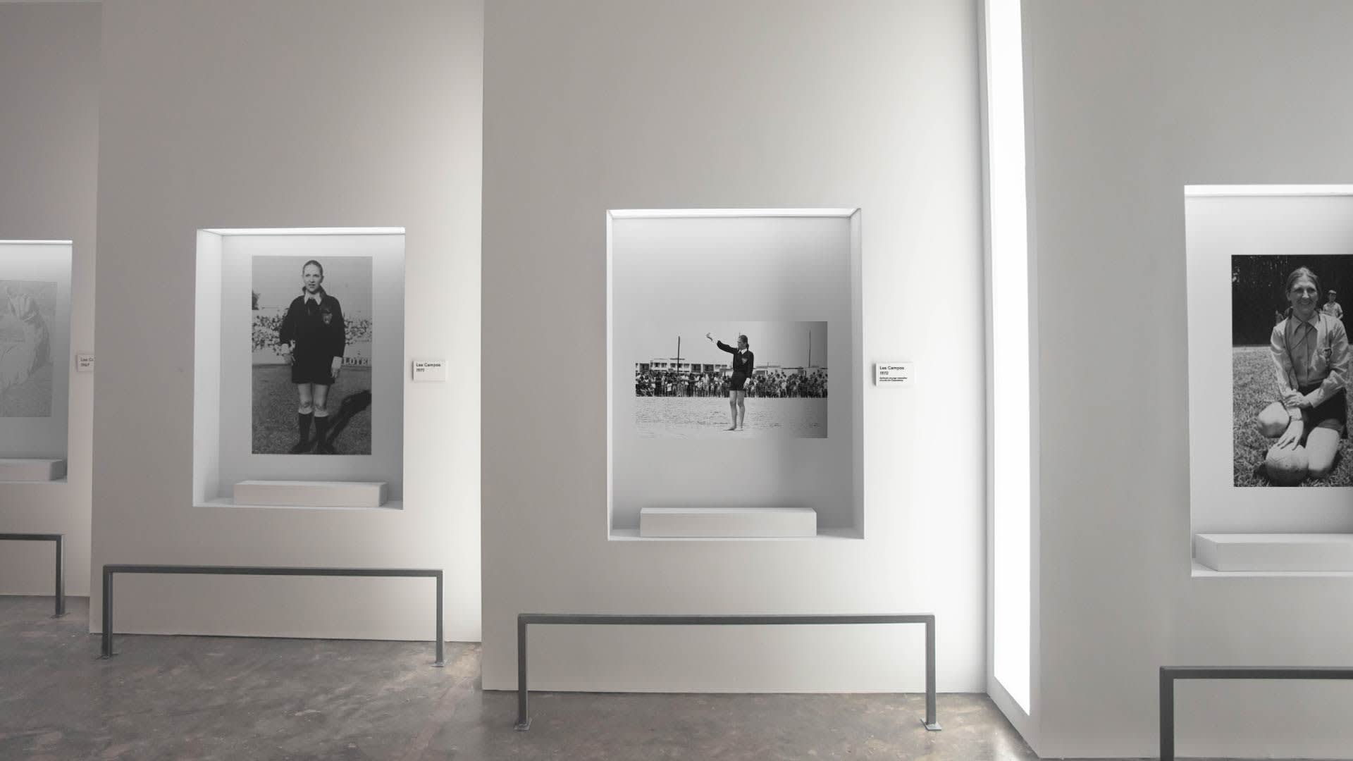 A modern art gallery with white walls displays three black-and-white photographs, each set in its own recessed, well-lit frame, depicting people in different outdoor scenes. Benches are placed below the displays.