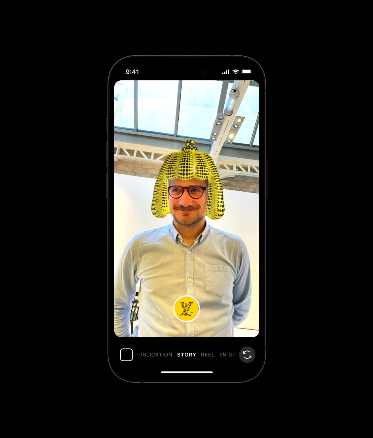 A man wearing glasses and a light blue shirt stands indoors. On a smartphone screen, an augmented reality filter adds a yellow and black Egyptian pharaoh headdress and a Louis Vuitton logo to his image.