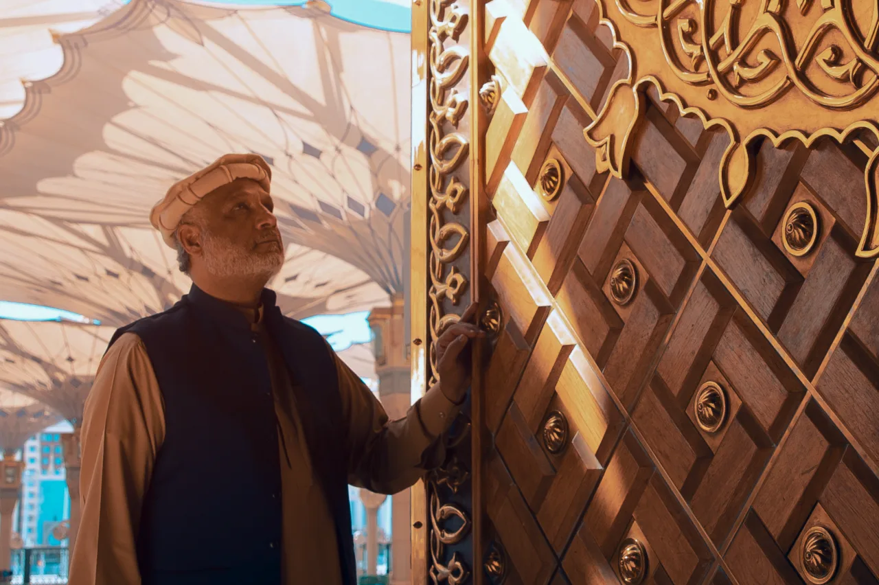 A man in traditional attire stands thoughtfully beside an ornate wooden door with intricate golden designs. Bright sunlight filters through large, patterned canopies in the background.