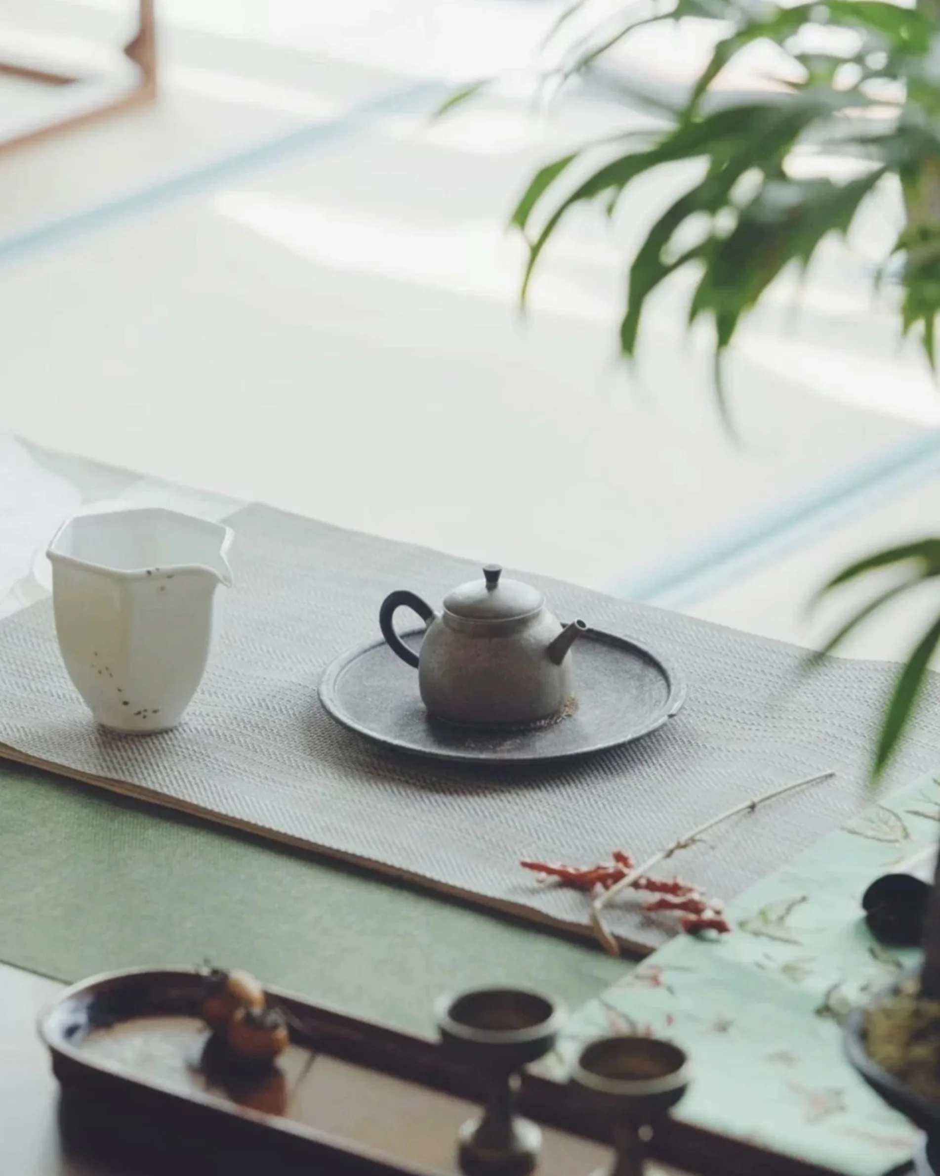 A small teapot sits on a round tray next to a white pitcher on a placemat, with cups and decorative plants nearby, creating a serene tea setting in soft, natural light.