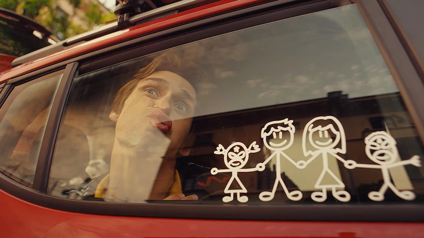 A man presses his face comically against the window of a red car. The window displays white stick-figure drawings of a family: two adults and two children holding hands, with one child making a silly face.