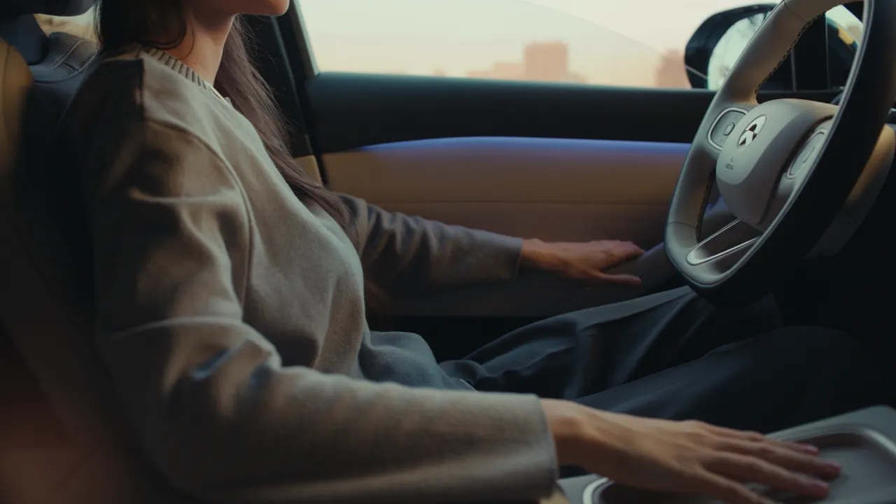 A person with long hair sits in the drivers seat of a car, resting one hand on the steering wheel and the other on the center console. The interior appears modern, with city buildings visible through the window.
