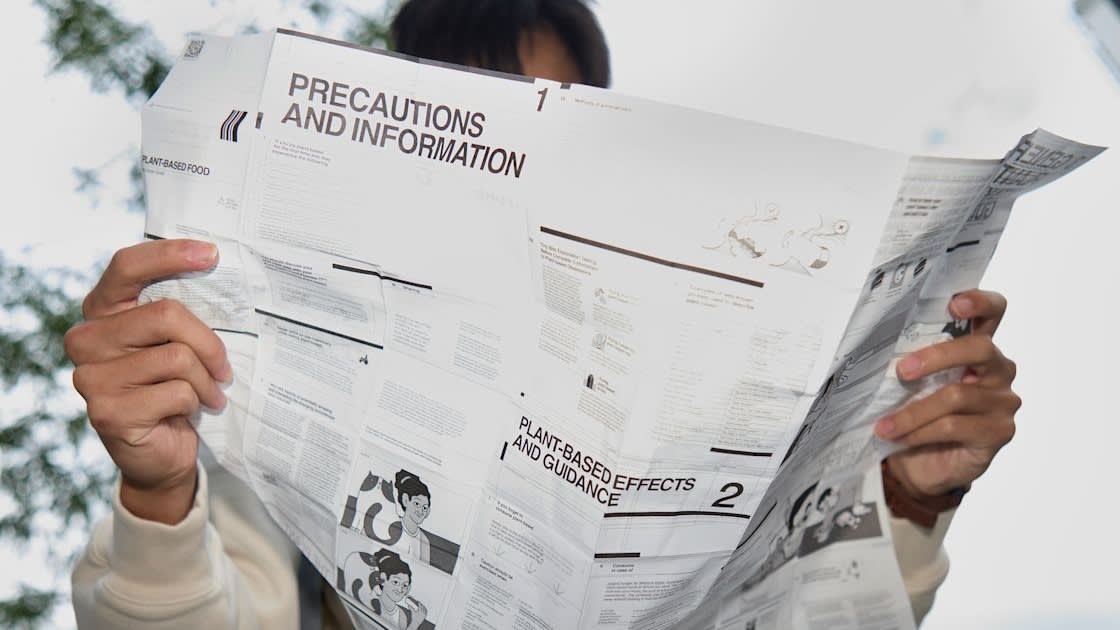 A person holds and reads a large pamphlet or instruction sheet that has the heading PRECAUTIONS AND INFORMATION, with some illustrations and text visible, but their face is mostly obscured.