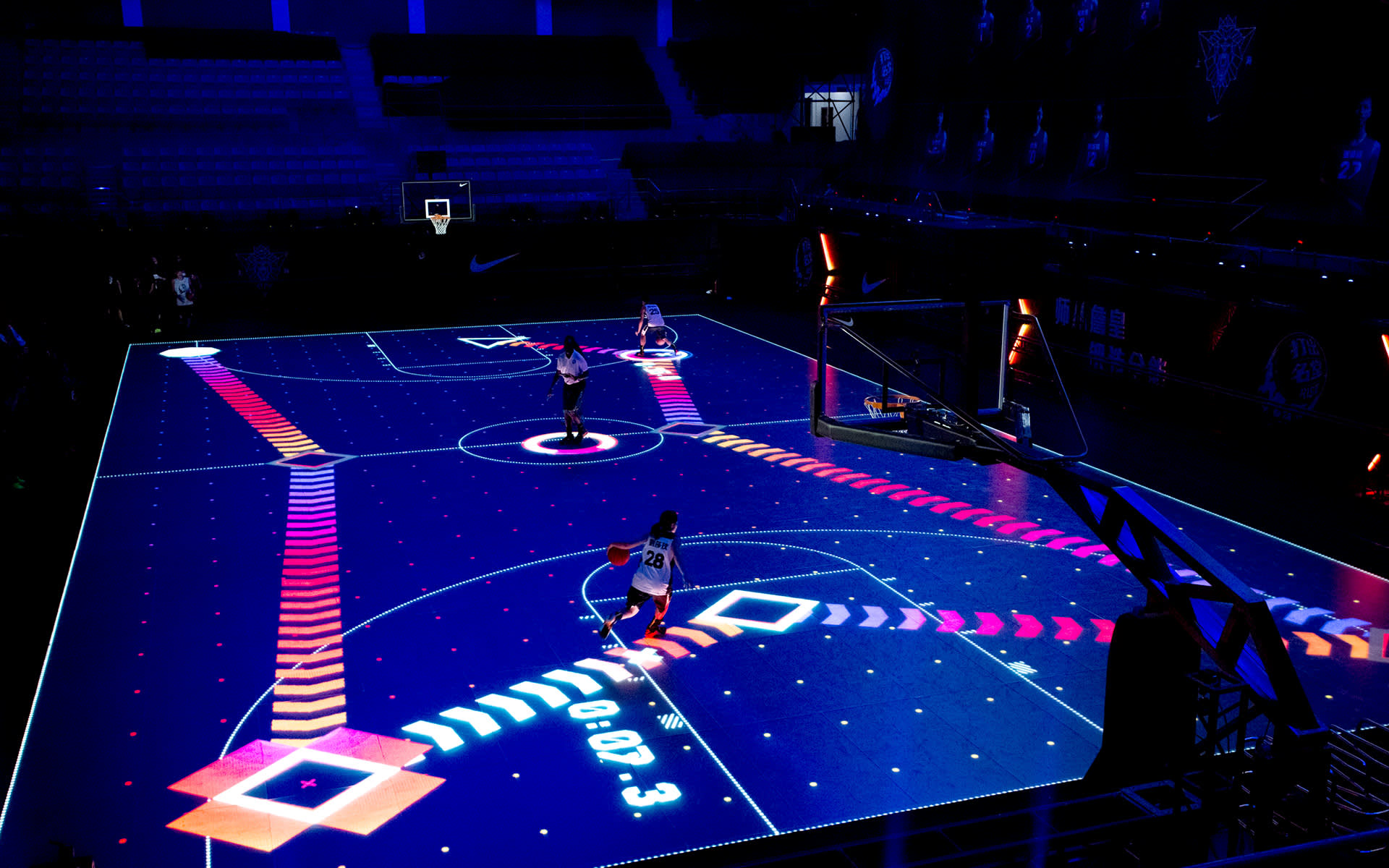Three basketball players on a high-tech court with illuminated digital graphics, including arrows and shapes, projected onto the floor. The arena is dimly lit, highlighting the vibrant visual effects on the playing surface.