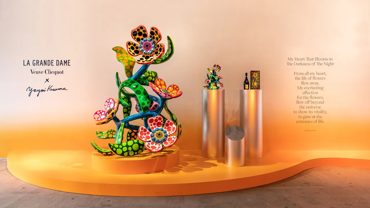 A vibrant, colorful floral sculpture stands on an orange platform beside silver cylindrical podiums displaying bottles. The wall features “La Grande Dame Veuve Clicquot x Yayoi Kusama” and a poetic inscription.