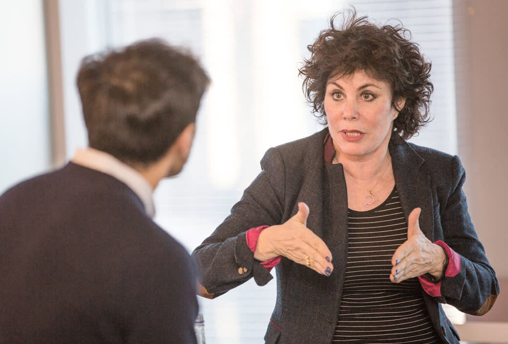 A woman with curly hair gestures with both hands while speaking to a man whose back is to the camera. They appear to be having an engaged conversation in a bright, indoor setting.