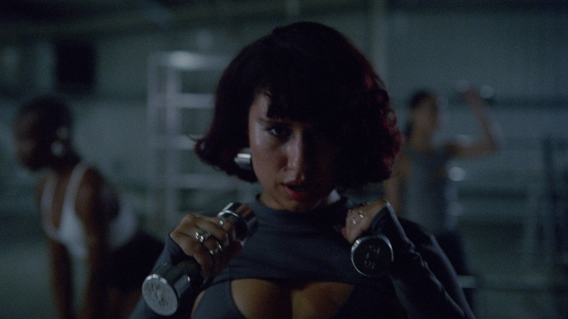 A woman with short dark hair and a cutout top holds two small dumbbells at shoulder height in a dimly lit gym. Two other people exercise in the blurry background.