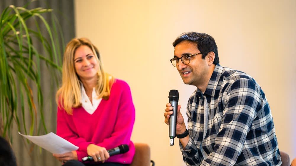 A man in a plaid shirt speaks into a microphone while sitting next to a smiling woman in a pink sweater who is holding papers. They appear to be participating in a casual discussion or interview. A plant is visible in the background.