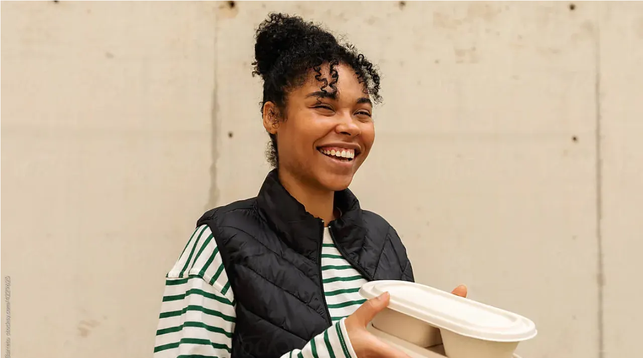 A smiling woman with curly hair, wearing a black vest over a green and white striped shirt, holds two compostable food containers against a plain light-colored background.