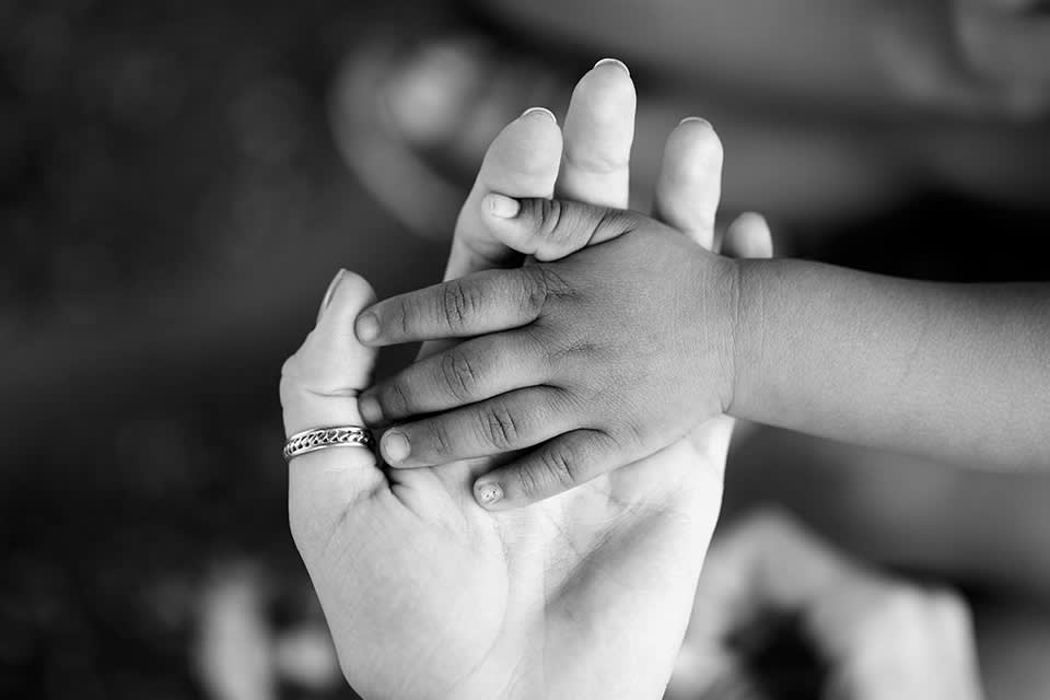 A black-and-white photo shows a childs hand resting gently on top of an adults open palm. The adult wears a ring, and the background is softly blurred.