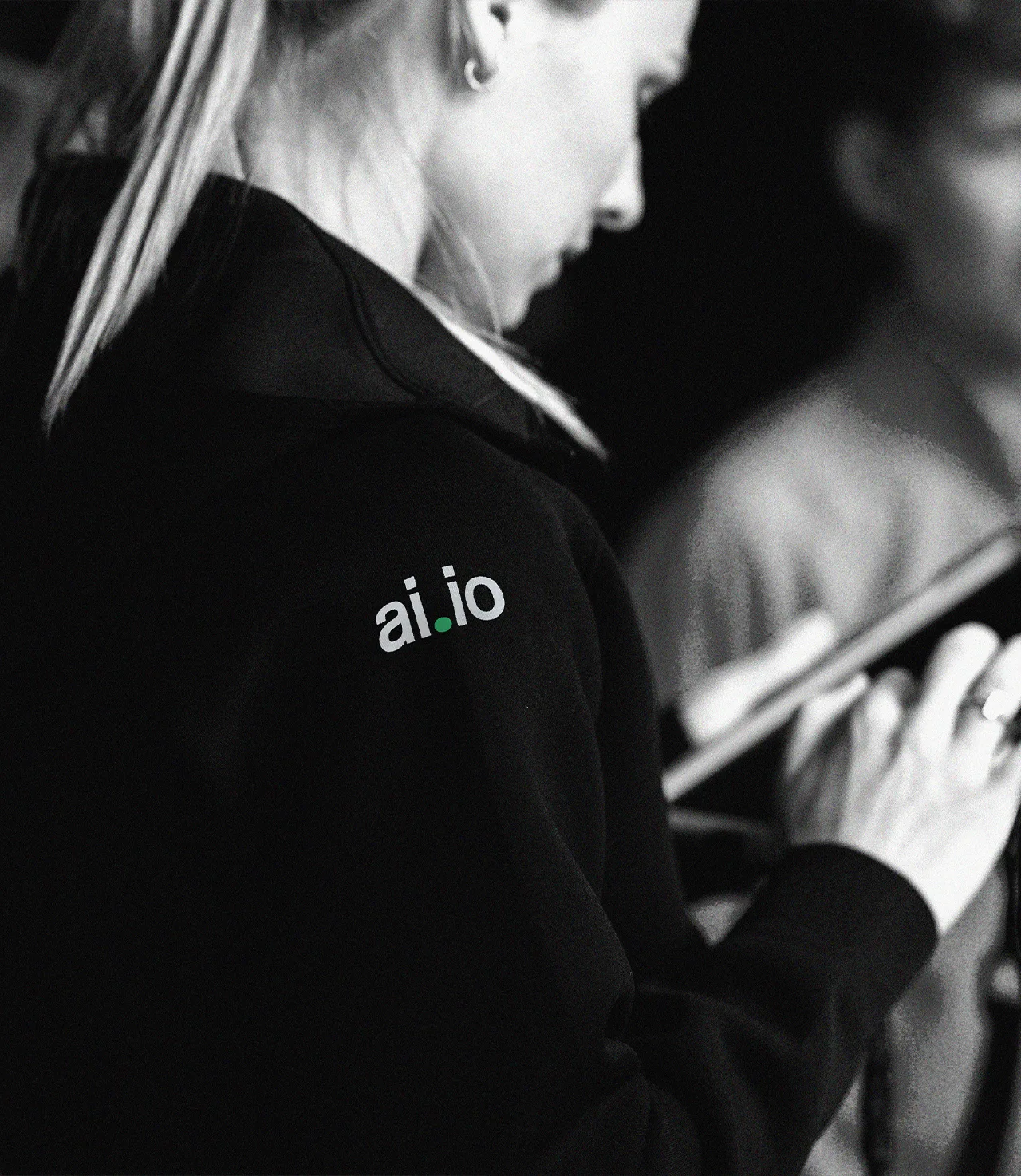 A person wearing a dark hoodie with the logo ai.io on the shoulder is holding and looking at a tablet. The image is in black and white.