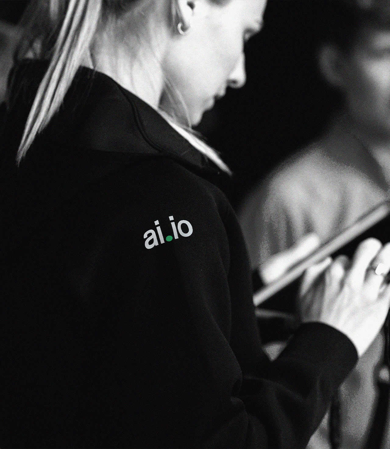 A person wearing a dark hoodie with the logo ai.io on the shoulder is holding and looking at a tablet. The image is in black and white.