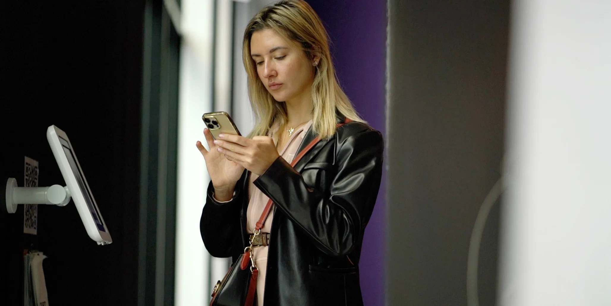 A woman with blonde hair wearing a black leather jacket stands indoors, looking at her smartphone and typing. She appears focused, with a crossbody bag and a neutral-colored top. A kiosk or stand is visible nearby.