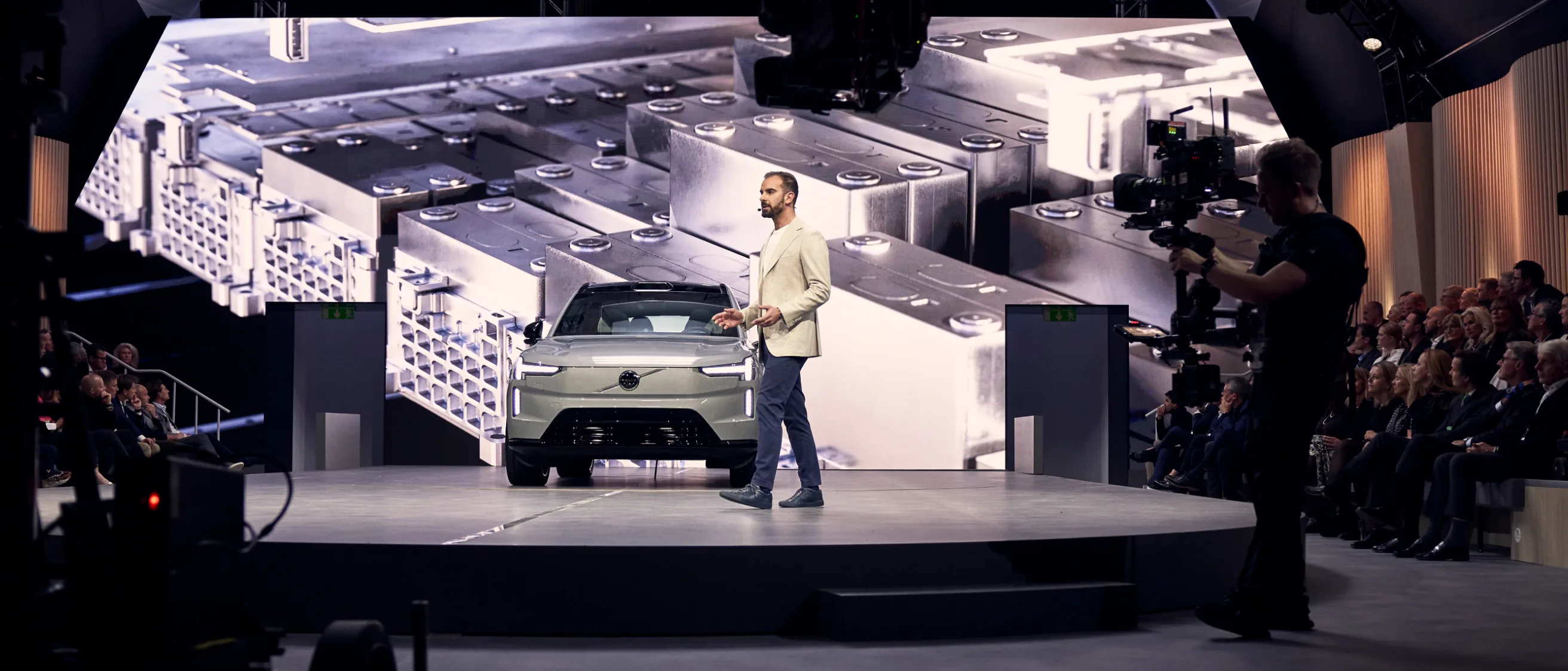 A man stands on a circular stage in front of an electric car and a large screen showing battery cells, while an audience and a camera operator observe the presentation.