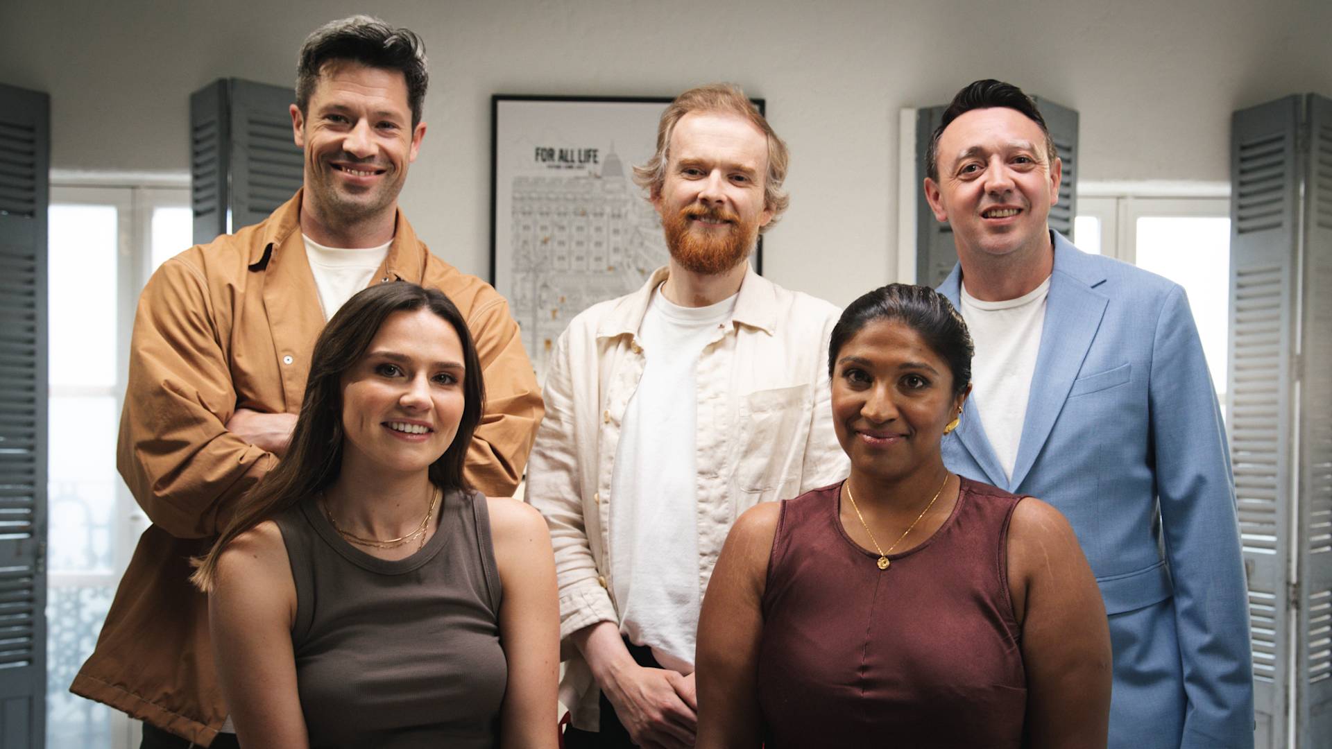 Five people pose and smile indoors in a well-lit room with grey shuttered windows and a framed poster on the wall behind them. Three stand in the back and two sit in front.