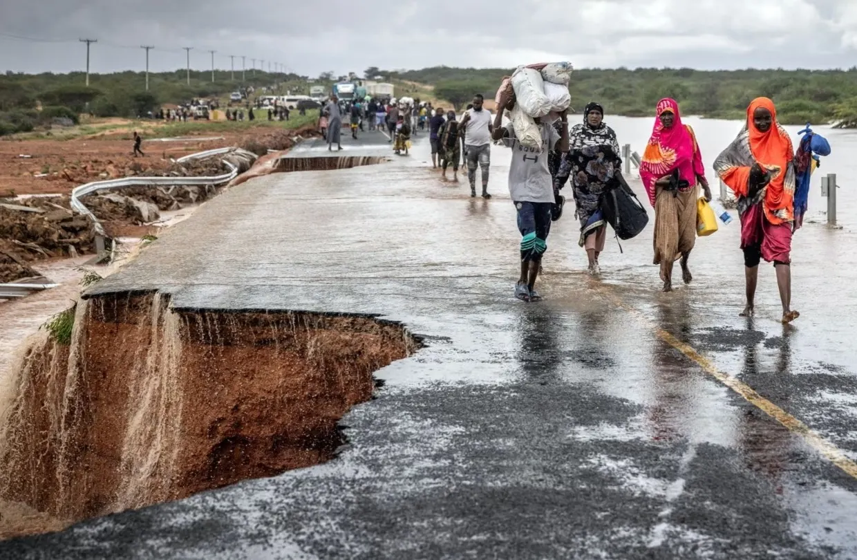 People walk along a rain-soaked, damaged road with a large section washed away by flooding; some carry belongings, and the sky is overcast, with more people in the distance.