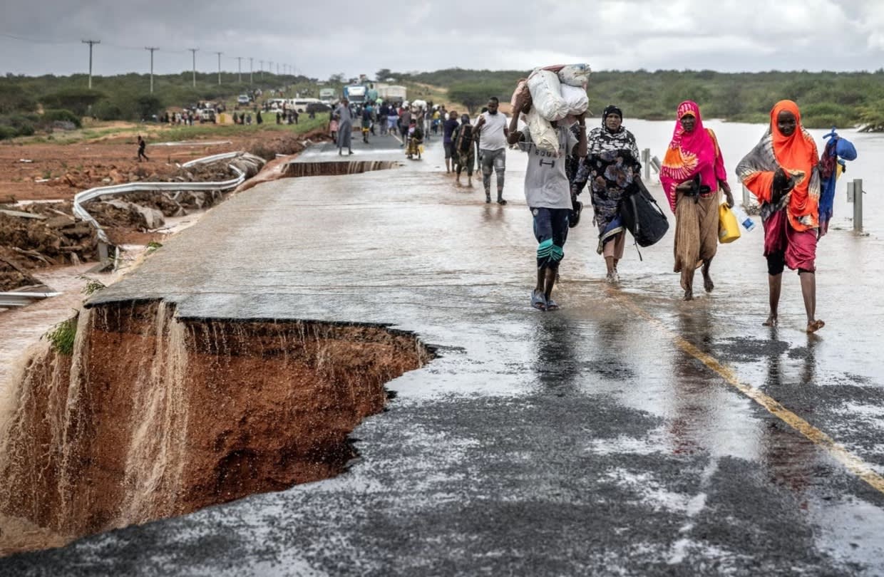 People walk along a rain-soaked, damaged road with a large section washed away by flooding; some carry belongings, and the sky is overcast, with more people in the distance.