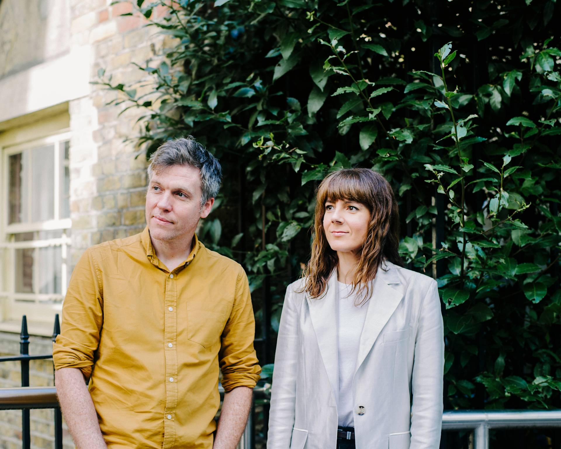 A man in a mustard yellow shirt and a woman in a light-colored blazer stand side by side outdoors, in front of leafy green bushes and a brick wall, looking slightly to their left.