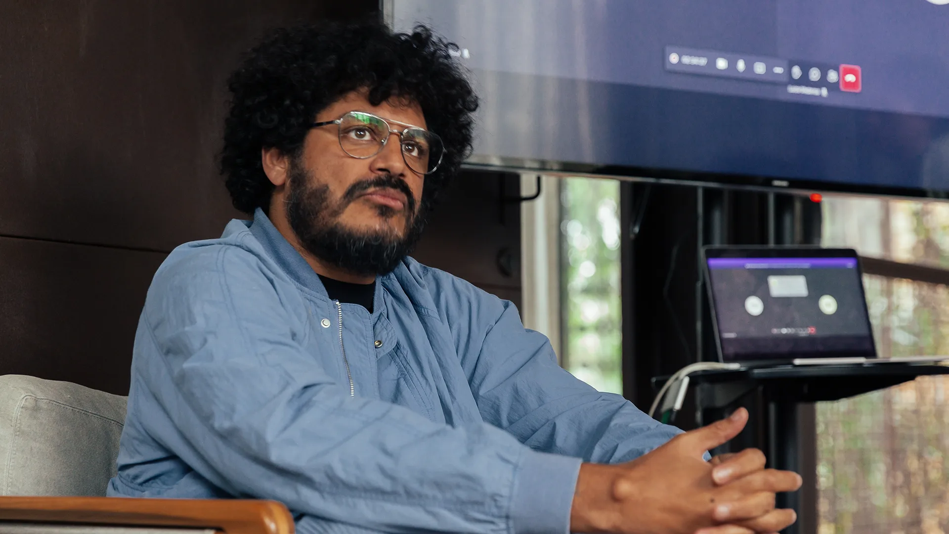 A man with curly hair, glasses, and a beard sits in a chair with clasped hands. He wears a blue jacket and looks thoughtful. Behind him are a large screen and a laptop on a stand.