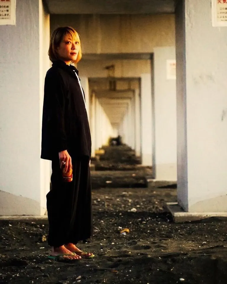 A person with short hair, wearing a loose black outfit and flip-flops, stands under a series of concrete arches with evening sunlight casting a warm glow. The ground is sandy and scattered with small debris.