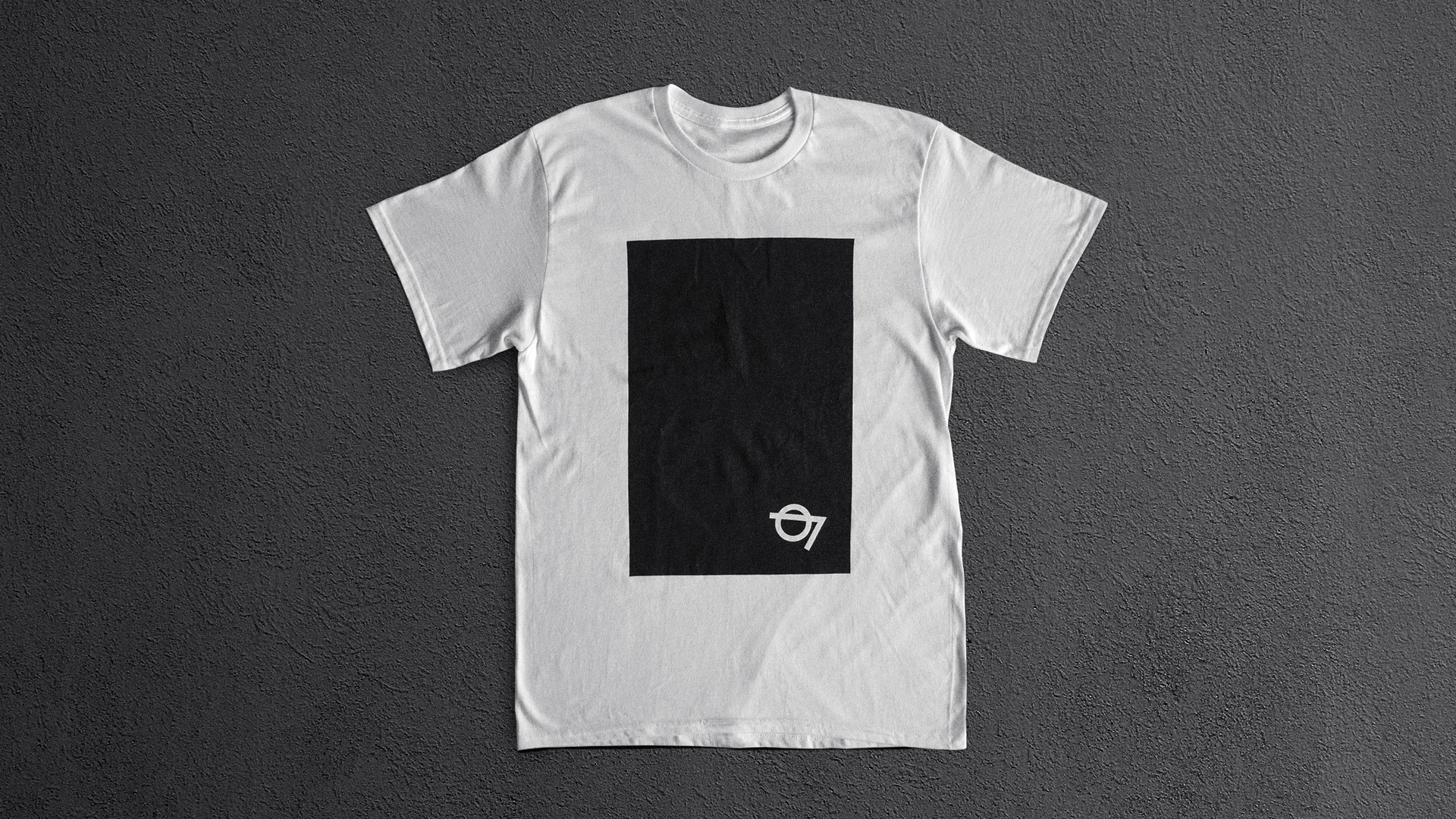 A white t-shirt laid flat on a gray textured surface, featuring a large black rectangle in the center and a small white 07 with a circle design in the lower right corner of the rectangle.