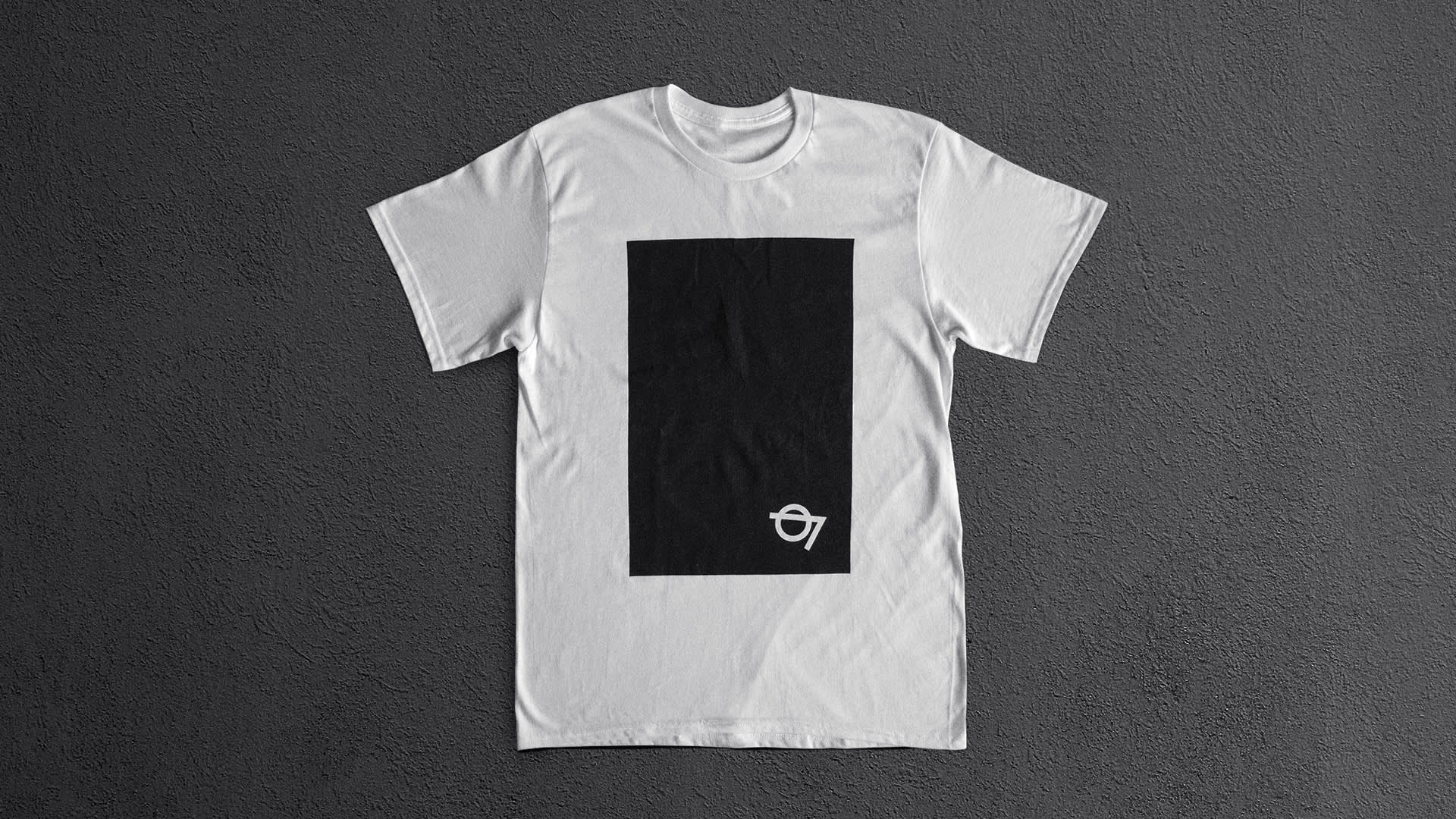 A white t-shirt laid flat on a gray textured surface, featuring a large black rectangle in the center and a small white 07 with a circle design in the lower right corner of the rectangle.