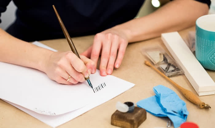 A person practices calligraphy on white paper using a dip pen. Various calligraphy tools, a turquoise mug, and blue paper are on the table nearby.