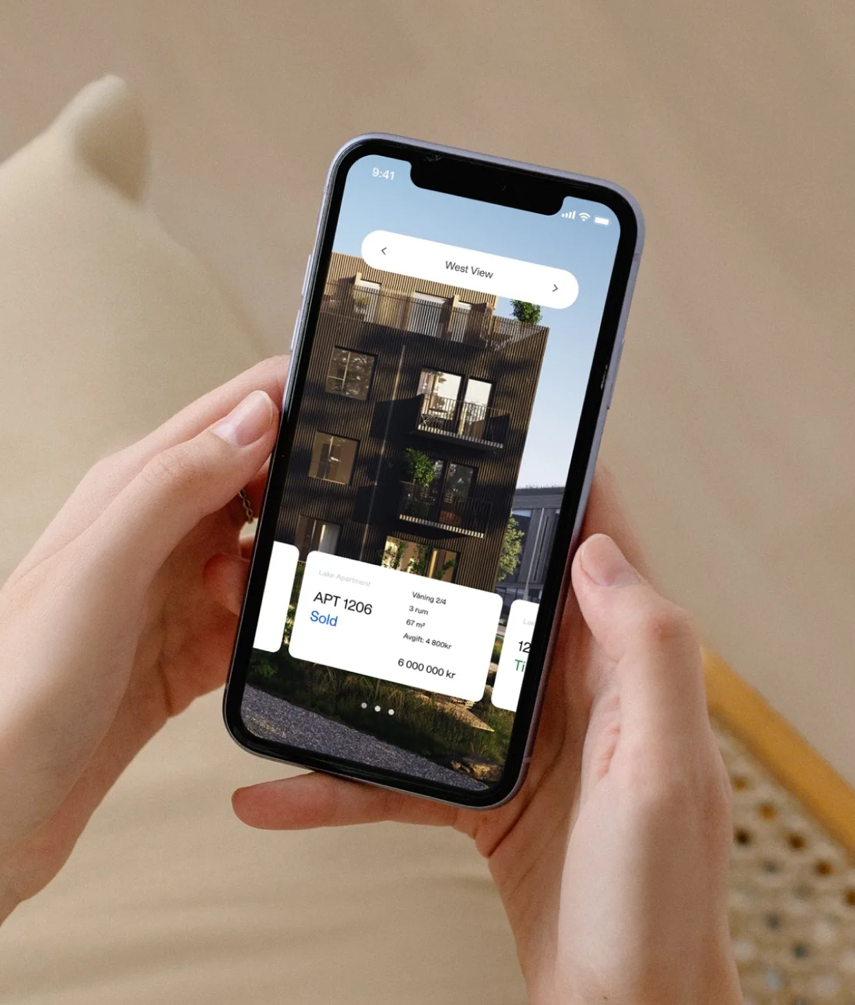 A person holds a smartphone displaying a real estate app with a photo of a modern apartment building and listings, including one that says “APT 1206 Sold.”.