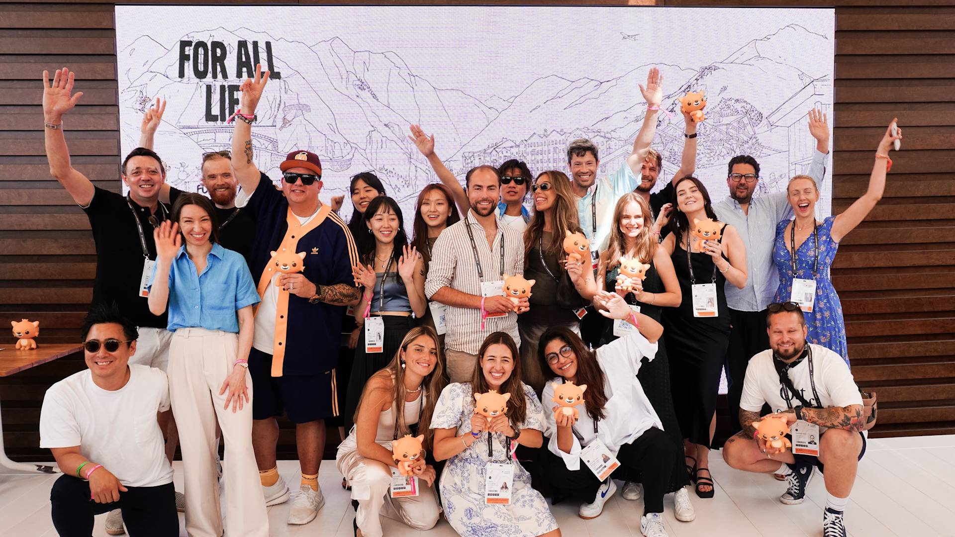 A diverse group of people smile and wave at the camera while holding small pig-shaped trophies, standing in front of a mountain-themed backdrop that reads FOR ALL LIFE.