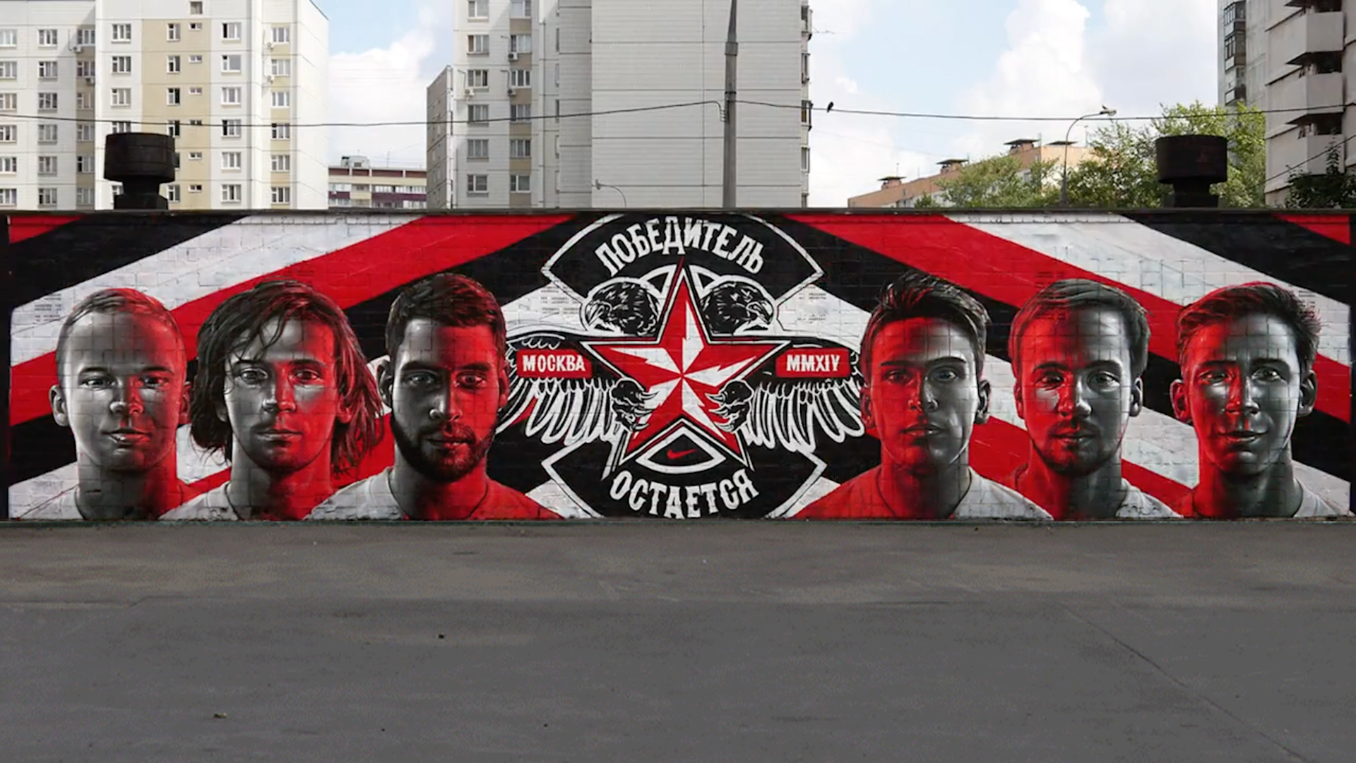 A mural features five serious-faced men in grayscale with red highlights, a red star with wings, and Russian text on a city wall, with apartment buildings visible in the background.