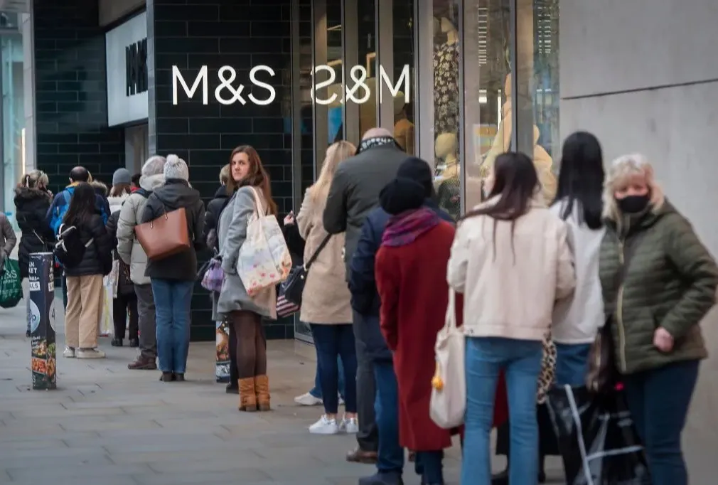 People wearing coats and scarves stand in a line outside an M&S store on a city street. Most face forward, some carry shopping bags, and a few wear masks. The shops glass windows display items inside.