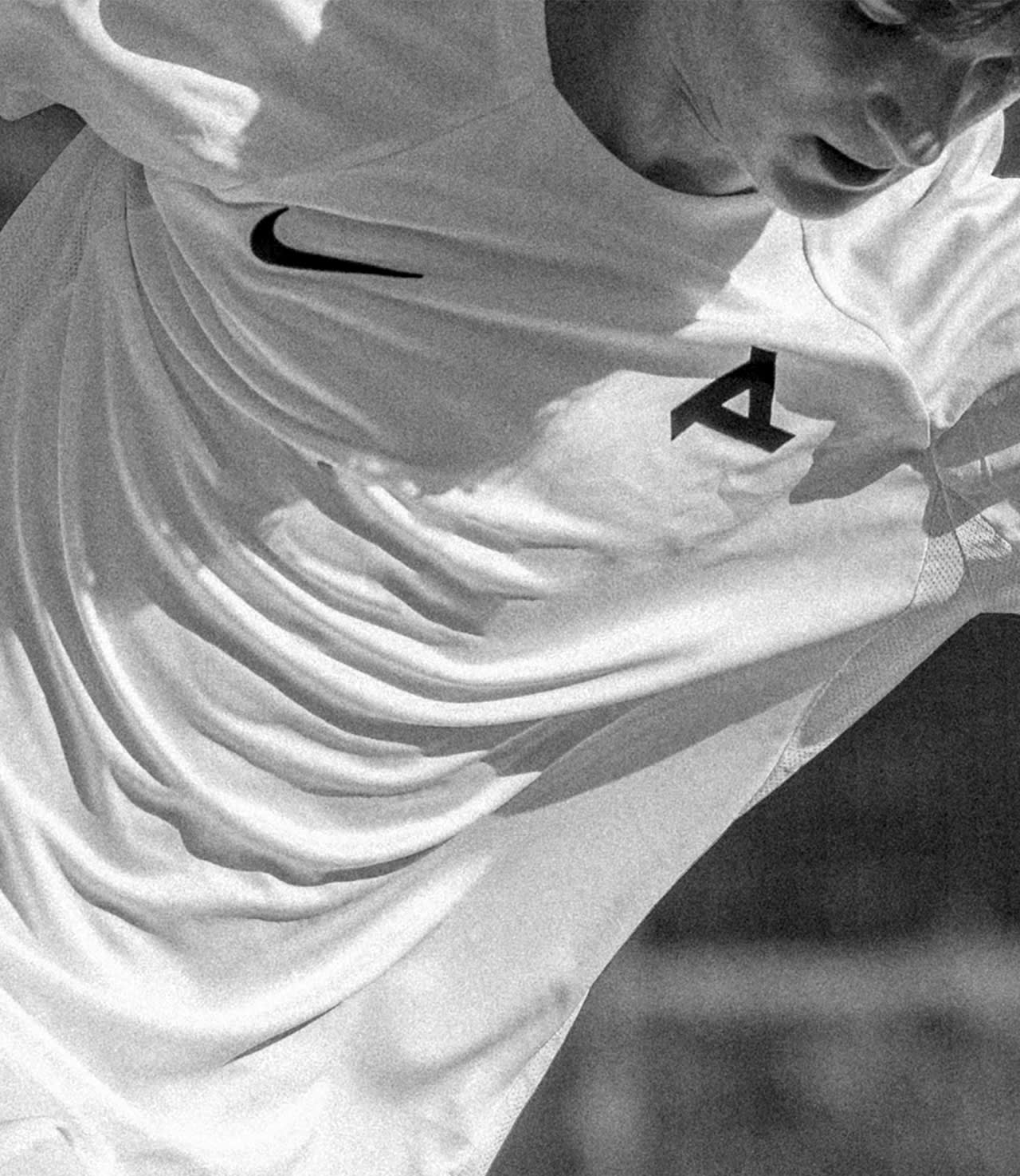 Black and white close-up of a person in a sports jersey, pulling the shirt at the chest. The jersey features a large letter A and a Nike logo. The persons face is partially visible.
