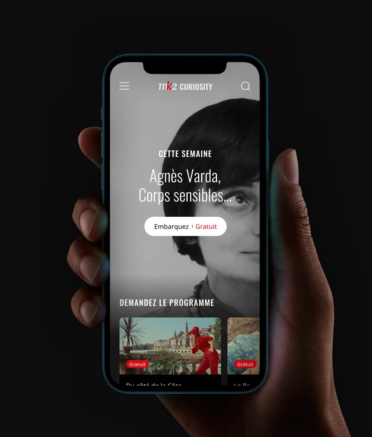 A hand holds a smartphone displaying the mk2 Curiosity app. The screen shows a black-and-white portrait of a woman and French text featuring Agnès Varda, Corps sensibles, along with options to explore or access content.
