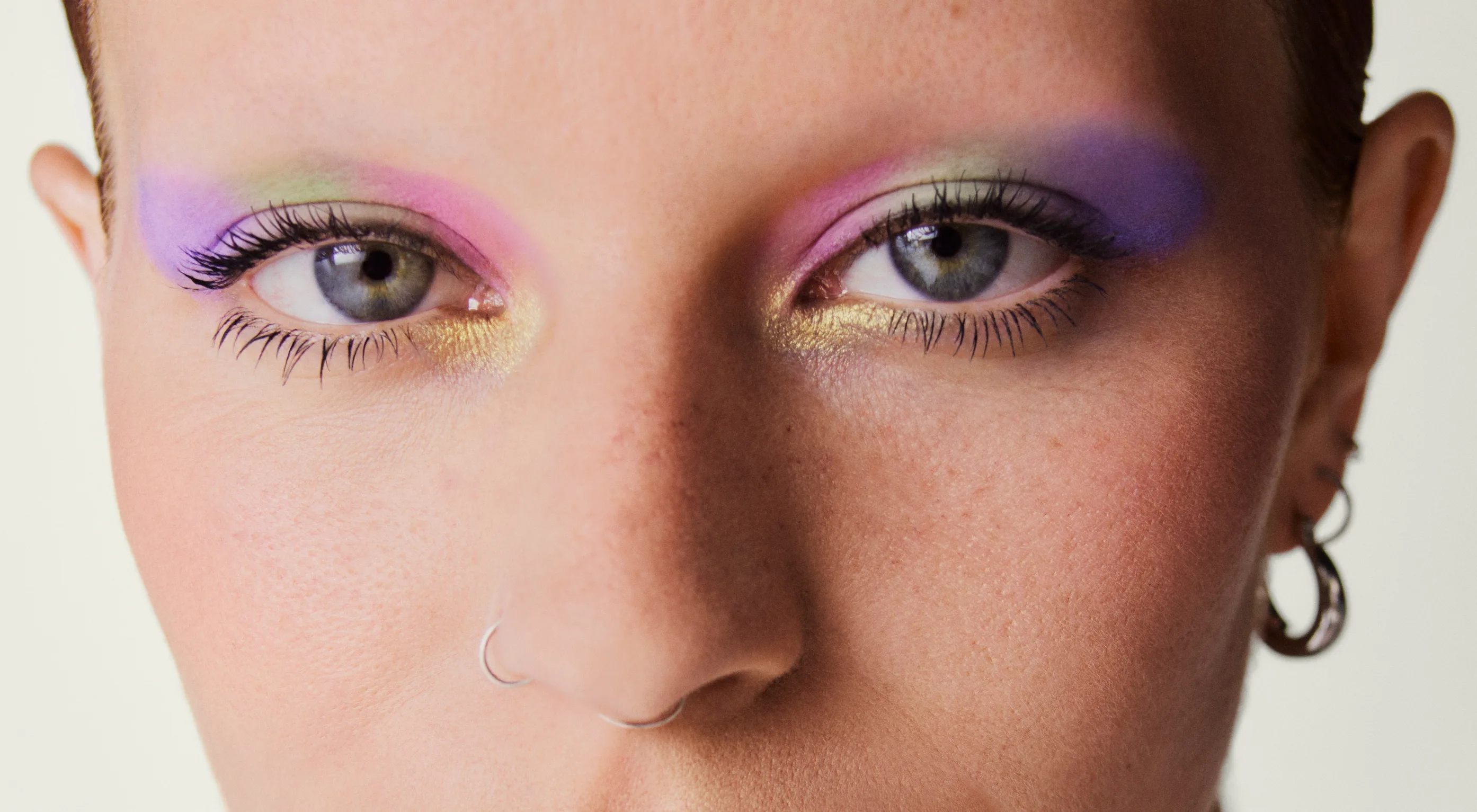 Close-up of a persons face with colorful pastel eye makeup in pink, green, purple, and gold shades, a silver nose ring, and a small hoop earring, with a neutral background.
