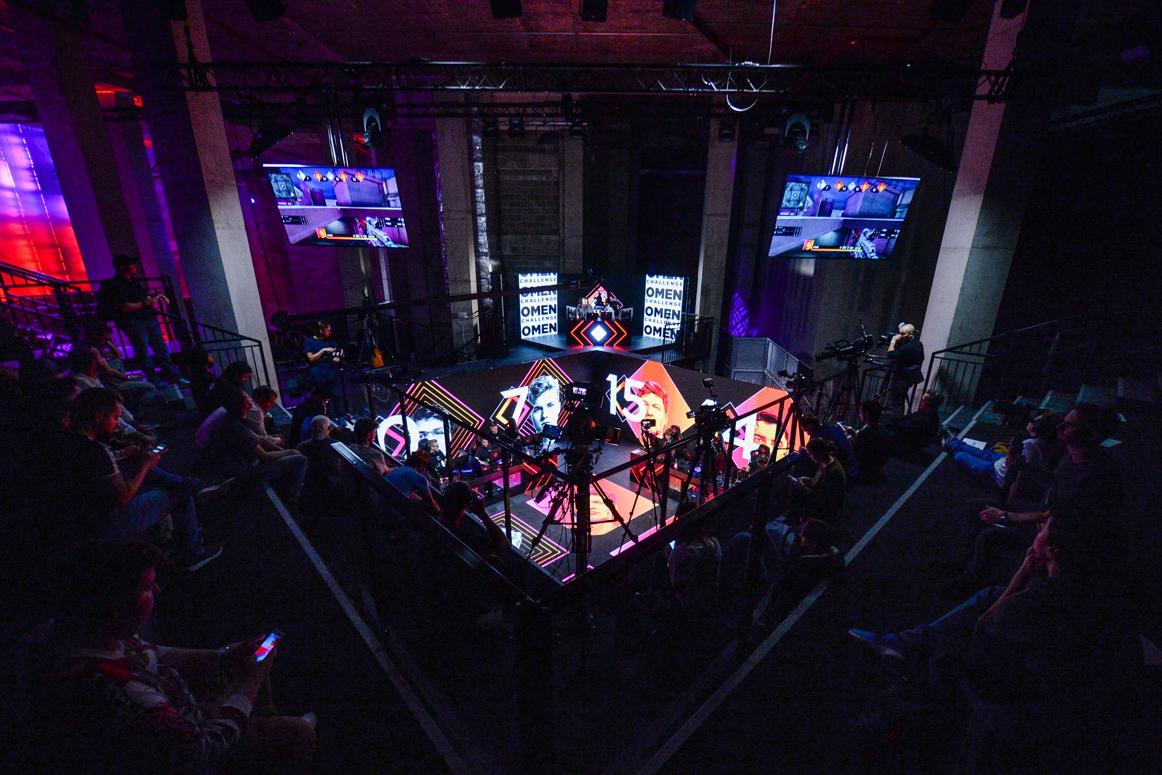 A gaming tournament stage with bright lights, multiple screens displaying gameplay, and an audience seated around the area. Several cameras and equipment surround the stage, capturing the event.