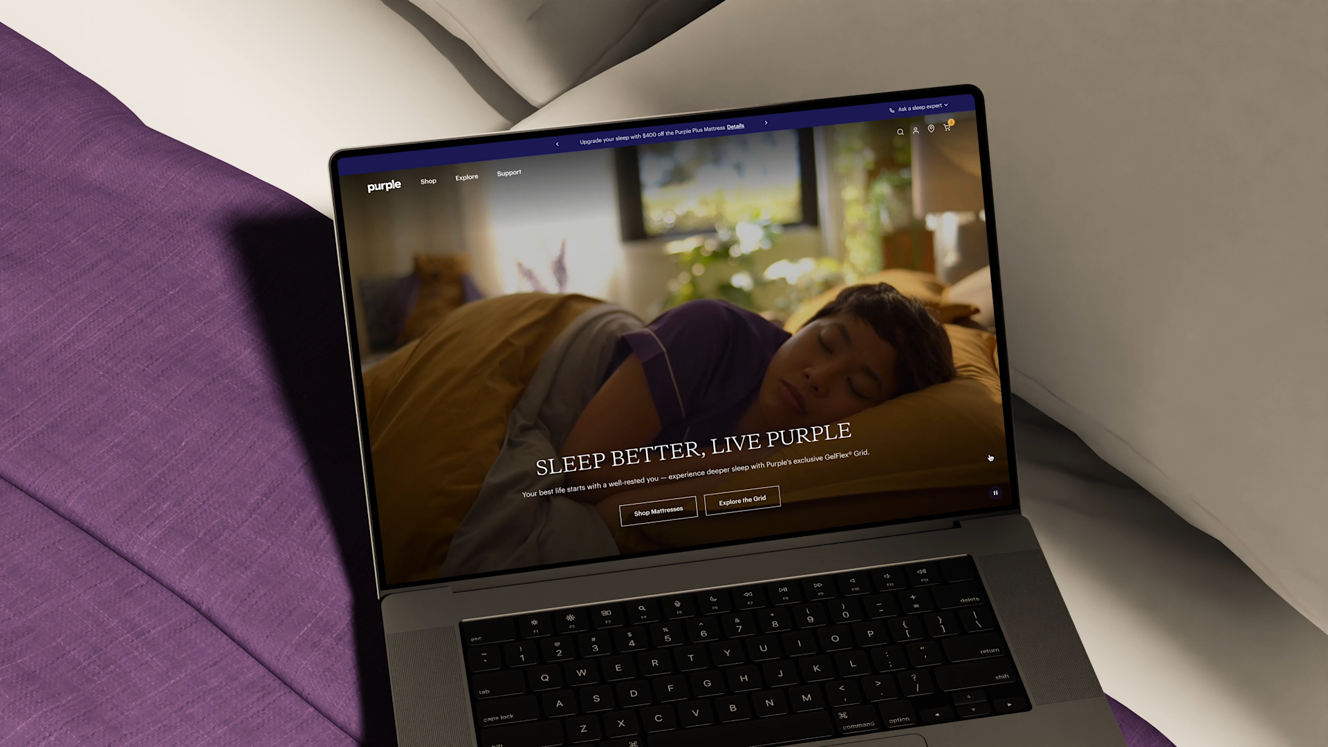 A laptop on a bed displays the Purple website, showing a person sleeping on their side under yellow bedding, with the headline Sleep Better, Live Purple. Purple sheets and pillows surround the laptop.