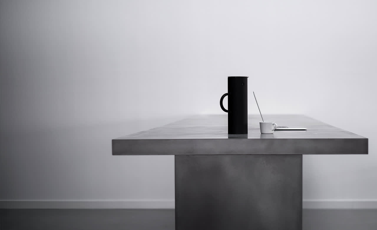 A minimalist scene with a long, gray table. On the table are a black thermos, a white cup with a spoon, and an open notebook, all arranged near the center. The background is plain and light-colored.
