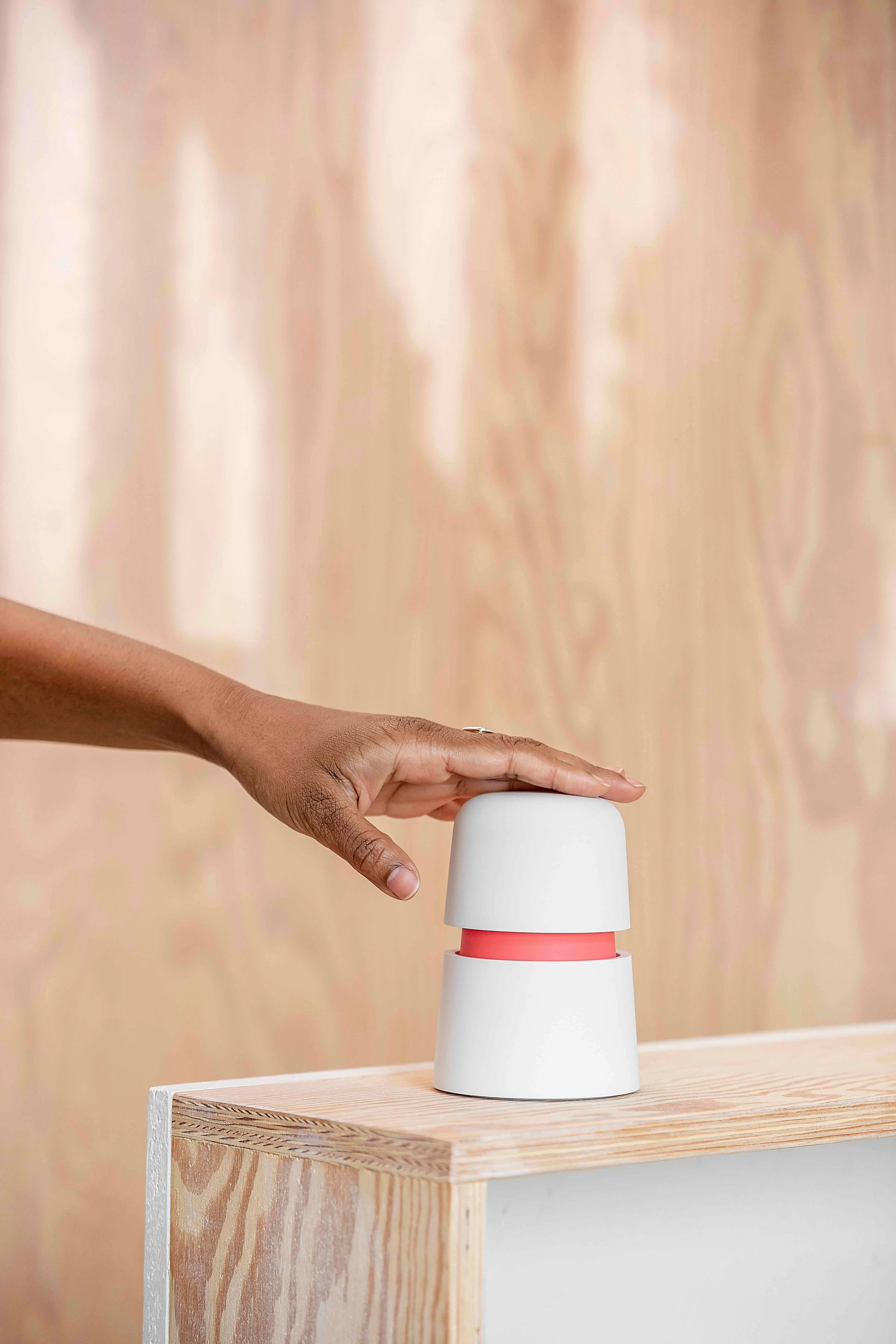 A hand pressing down on a small, white cylindrical device with a red band, placed on a light wooden surface with a blurred wooden background.
