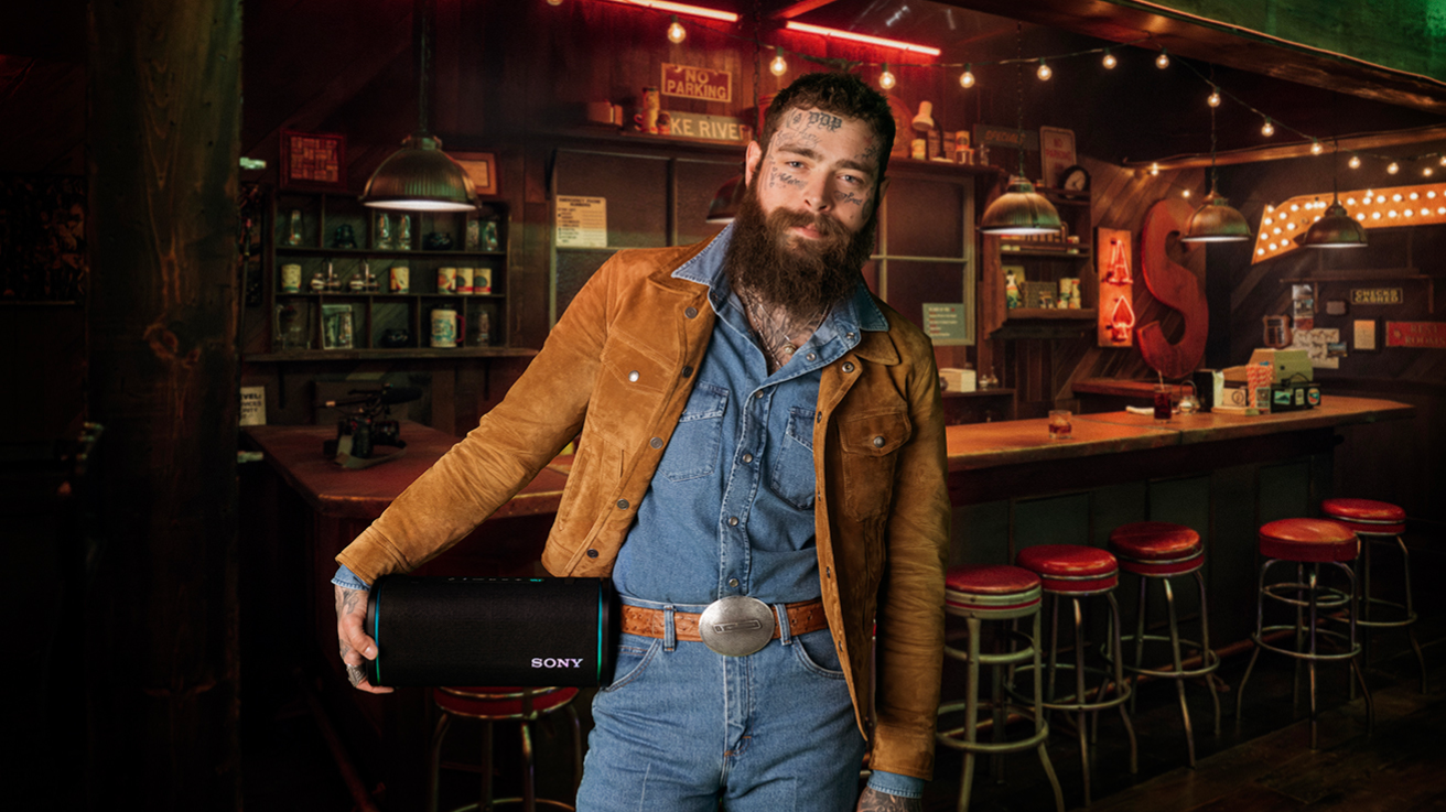 A bearded man in a brown suede jacket and denim outfit stands in a dimly lit bar, holding a black Sony speaker. Red bar stools and colorful lights are visible in the cozy background.