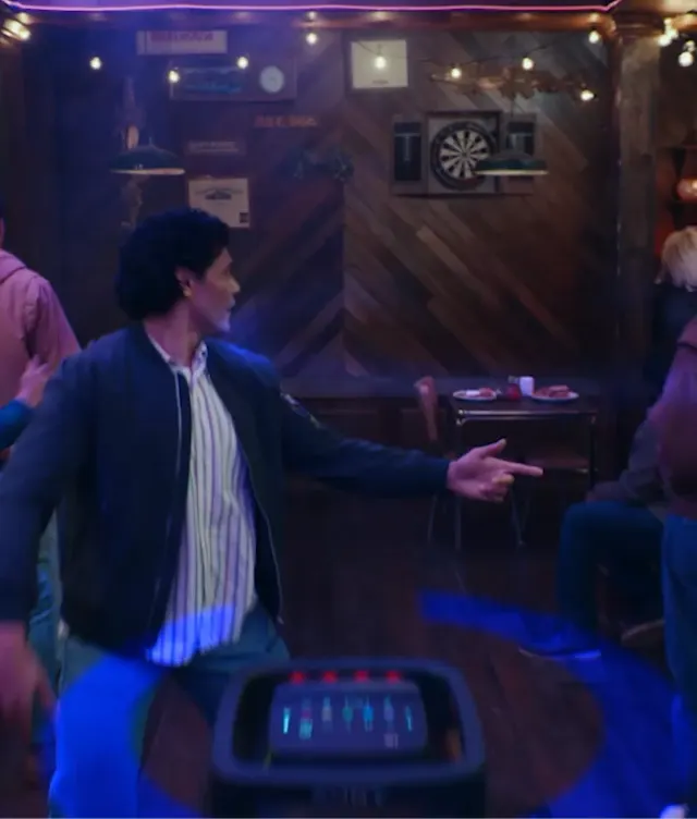 A man in a casual jacket and striped shirt stands in a dimly lit bar, pointing to his right. Several people are seated around him, with dartboards and wooden decor visible on the back wall.
