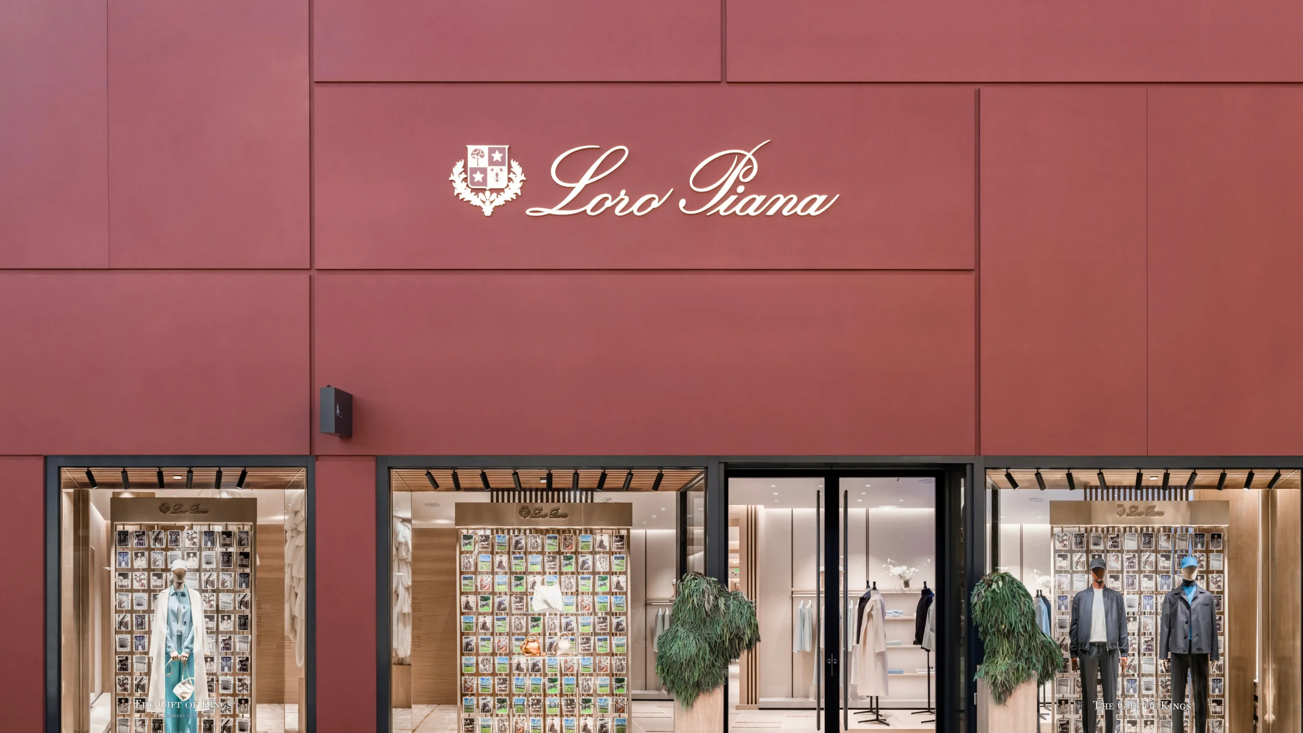 A storefront with a maroon facade features the Loro Piana logo in elegant white script above large display windows showing mannequins dressed in upscale clothing and a wall of neatly arranged colorful items inside.