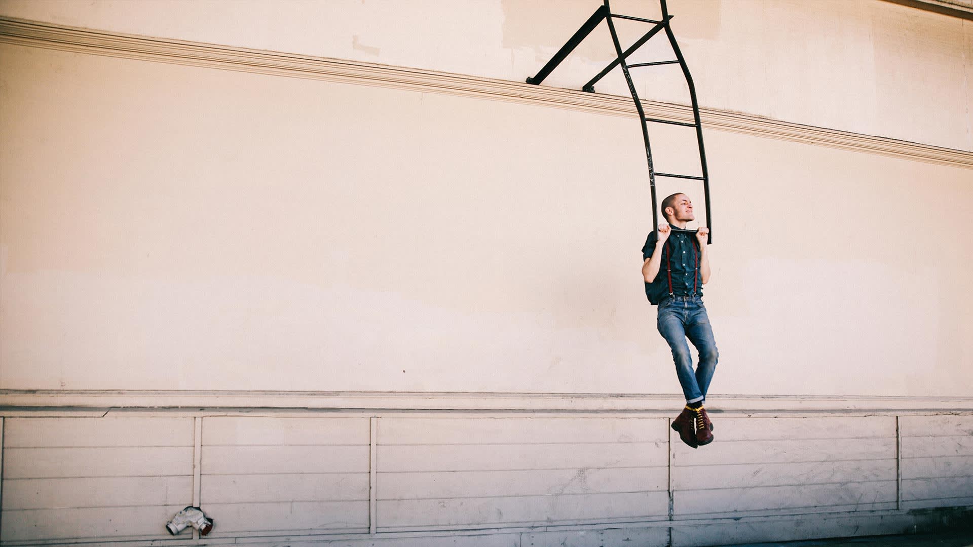 A person in jeans and a denim shirt hangs by their arms from a black ladder fixture attached to a beige wall, with feet crossed and the walls lower section featuring white paneling.