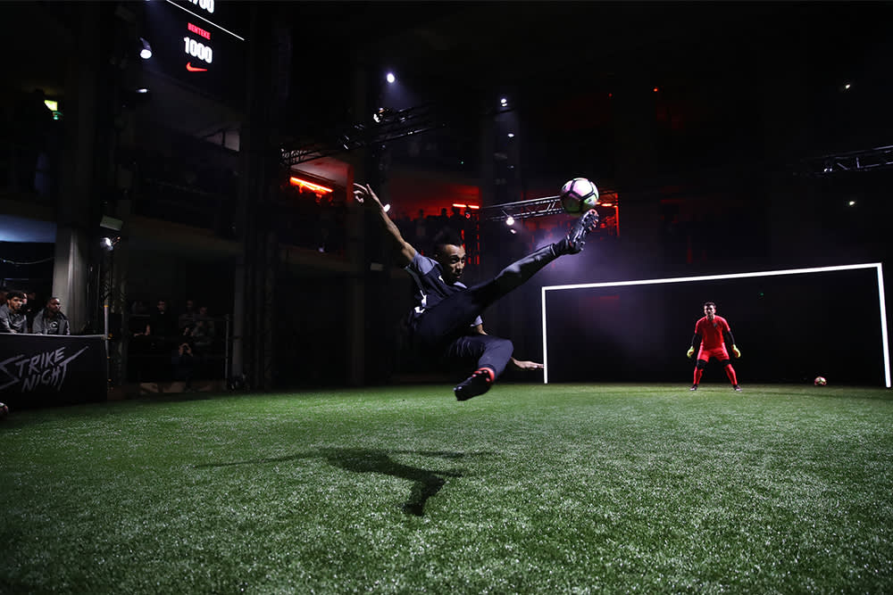 A soccer player performs an acrobatic kick in mid-air on an indoor field, while a goalkeeper in red stands ready in front of an illuminated goal. Spectators watch from the sidelines in a dark, dramatic setting.