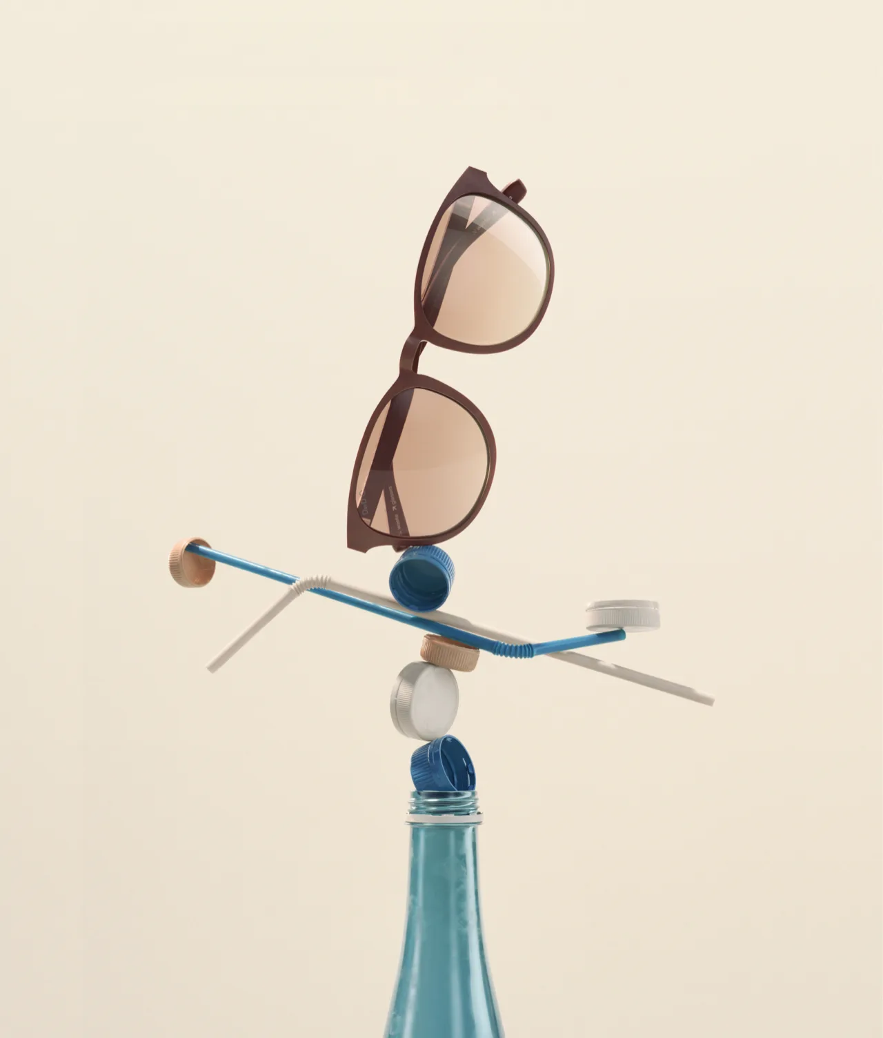 A pair of sunglasses balances on a stack of bottle caps, straws, and small objects, all precariously perched on the top of a blue glass bottle against a plain, light background.
