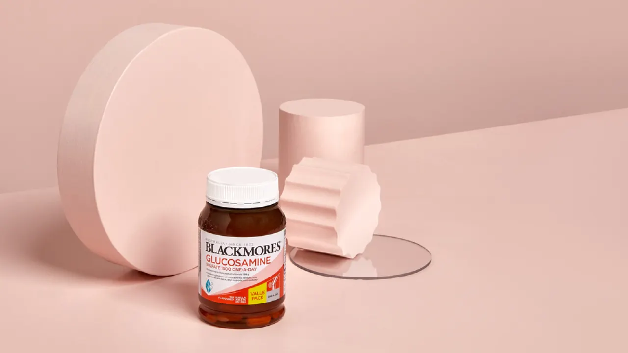 A jar of Blackmores Glucosamine is placed on a pale pink surface with light pink geometric shapes in the background. The scene has a minimalist, modern look.