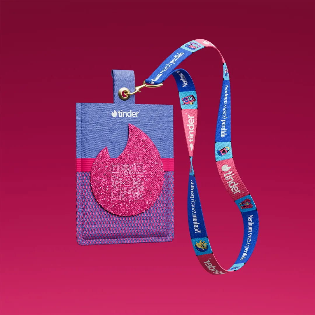 A blue badge holder with a pink glittery Tinder flame logo, attached to a blue and pink lanyard with Tinder branding and colorful icons, set against a gradient pink background.