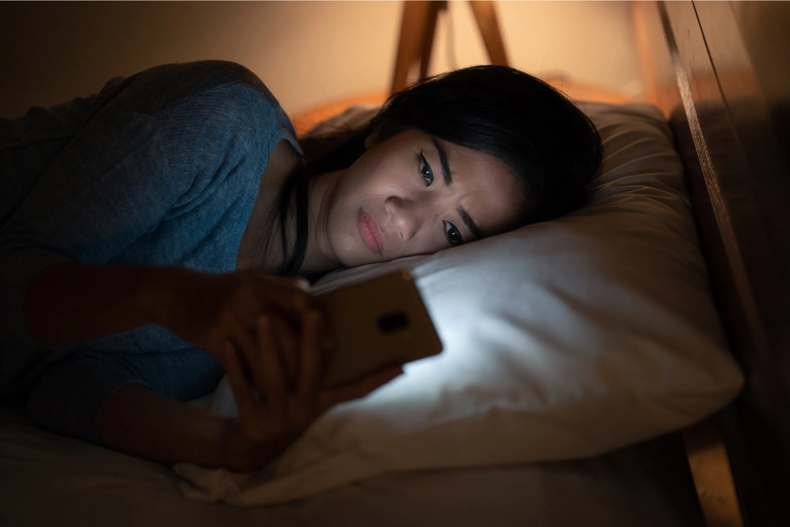 A woman lies in bed at night, looking at her smartphone with a concerned expression. The room is dimly lit, and the phone screen illuminates her face as she rests her head on a pillow.