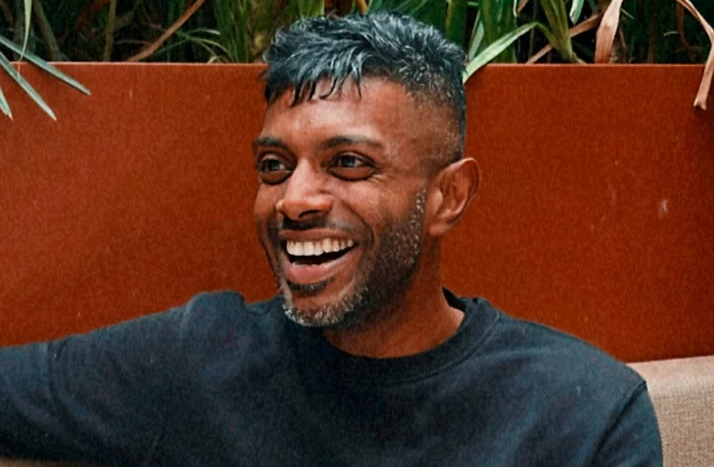 A man with short dark hair and a beard, wearing a black sweater, sits on a couch and smiles broadly. Green plants and a reddish-brown wall are in the background.
