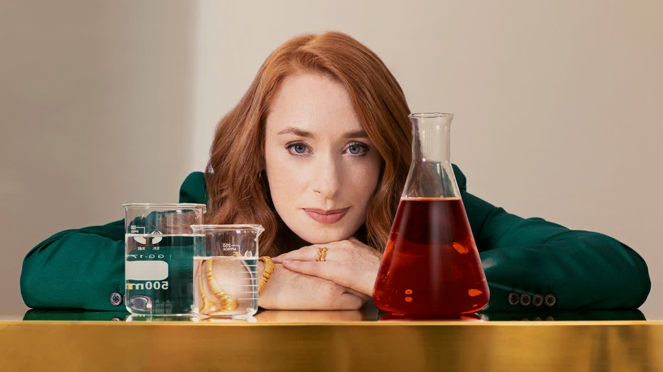 A woman with long red hair and a green blazer rests her chin on her hands, surrounded by three glass containers: two beakers with clear liquid and a worm, and a flask with a red liquid, on a reflective surface.